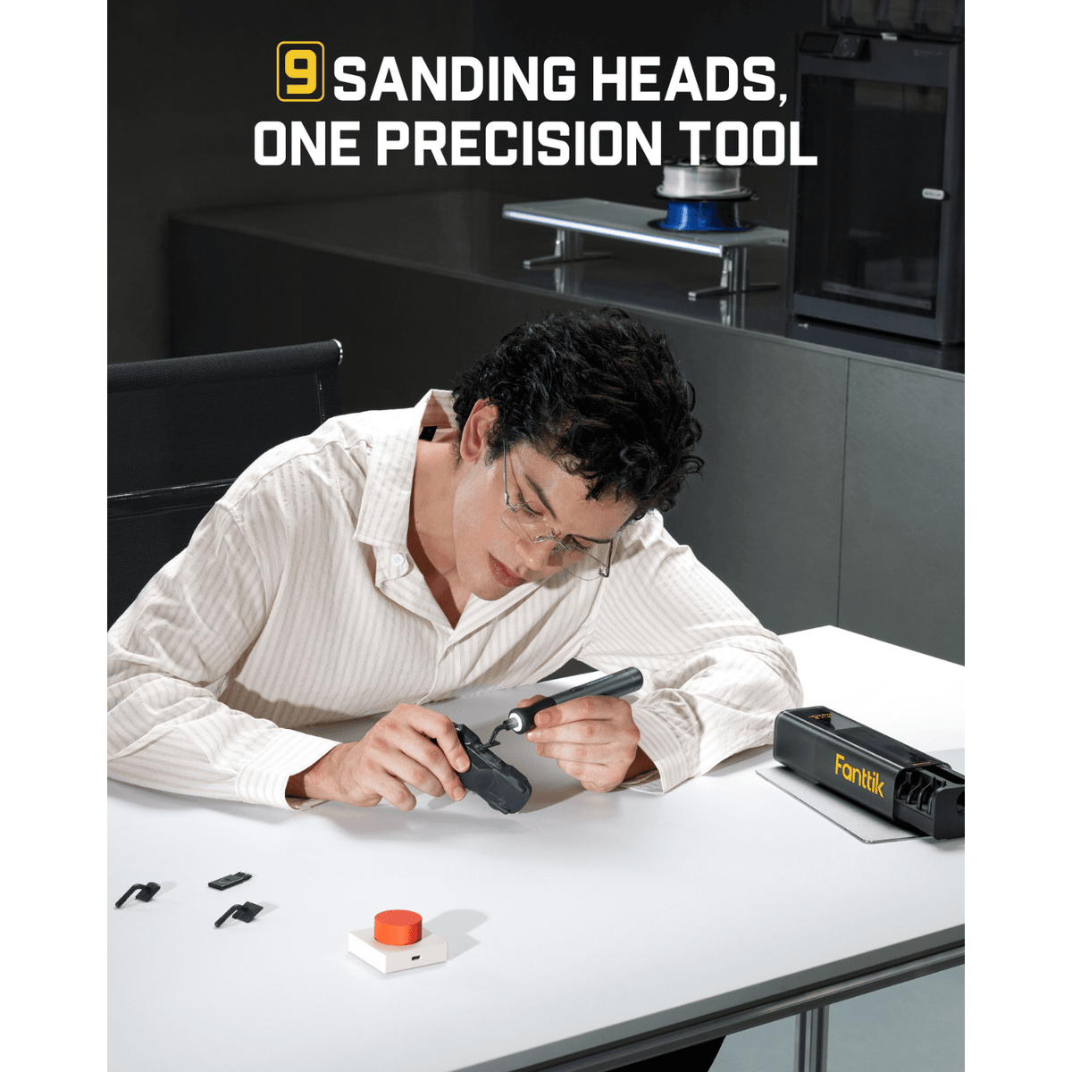 A man in a white shirt leans over a white table, using Fanttik Pix 3 Master Electric Detail Sanderon a small object. On the table are a black Fanttik-branded case, a small orange-capped container, and a few small black pieces.